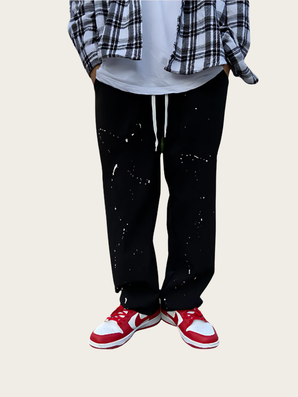 Person wearing black pants with white splatter design, plaid shirt, and red sneakers on a light background