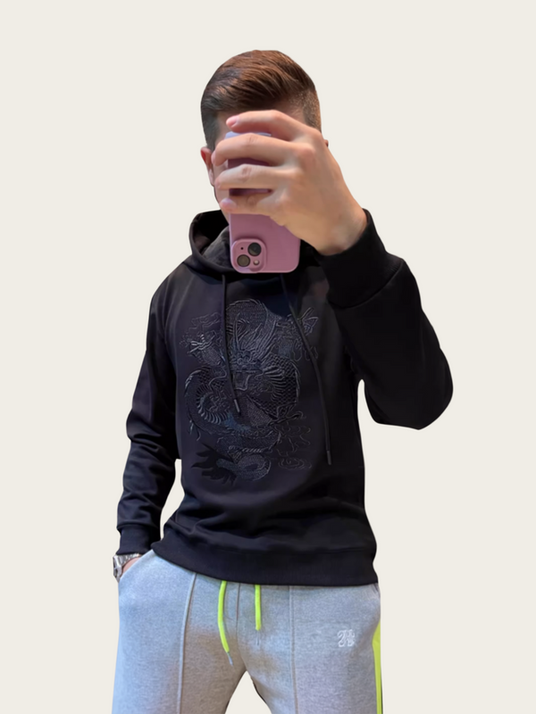 Person wearing a black hoodie with a design, taking a mirror selfie.