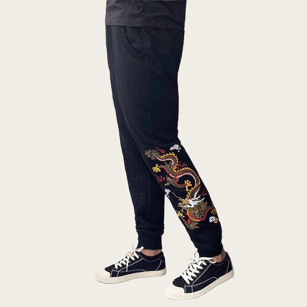 Black jogger pants with colorful dragon embroidery worn by a person on a white background