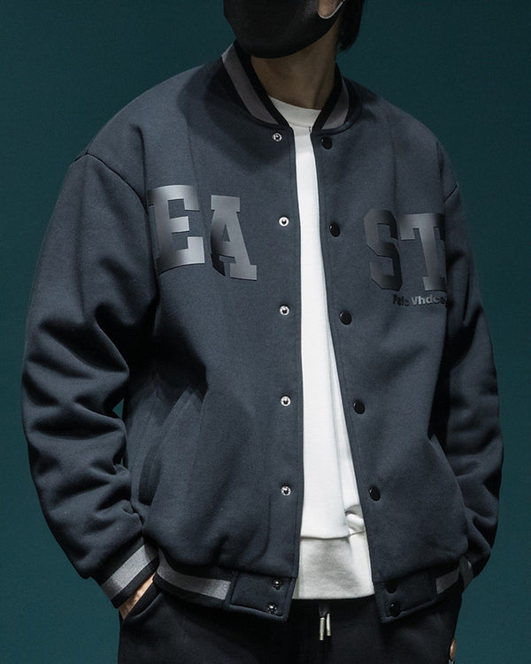 Dark slate-black varsity jacket with EA and ST chest letters, snap front and striped ribbed collar, worn over a white tee