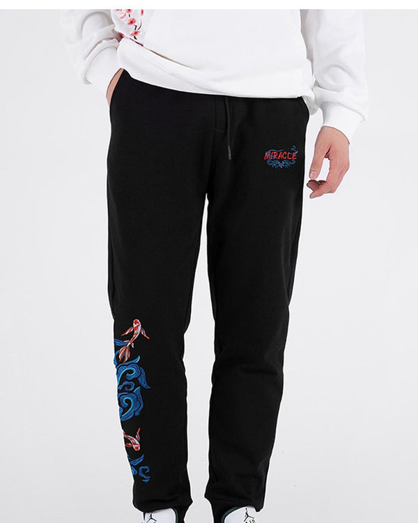 Man in black embroidered jogger pants with a white sweatshirt, front view with a hand in the pocket