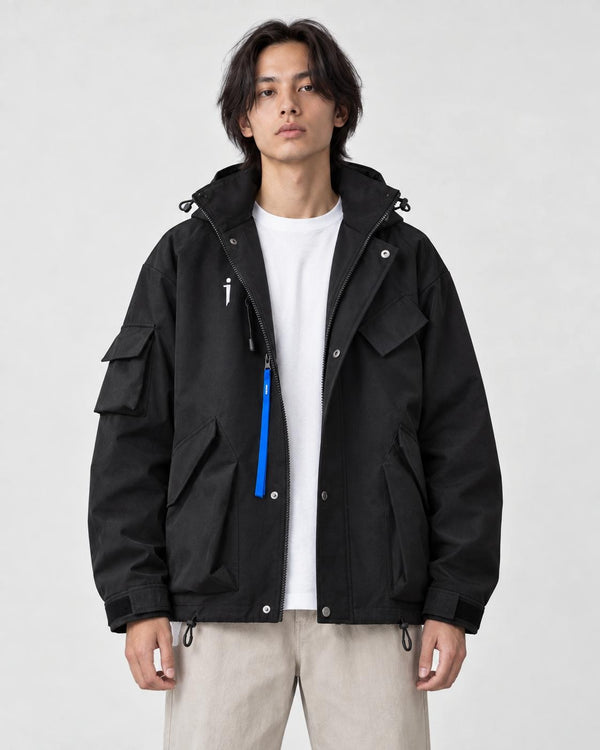 Man in a black hooded techwear jacket with a blue zip pull over a white tee and stone trousers, torso to mid-thigh