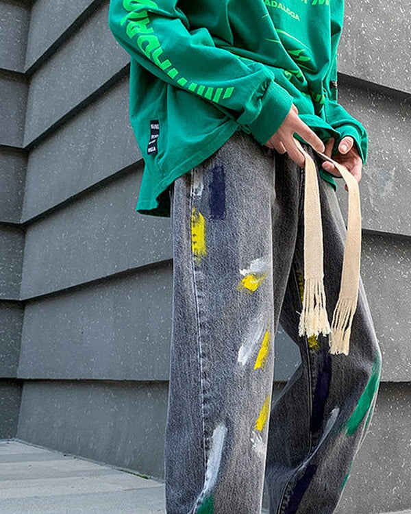 Washed gray paint-splatter jeans with two long frayed cotton tapes, worn with a green sweatshirt