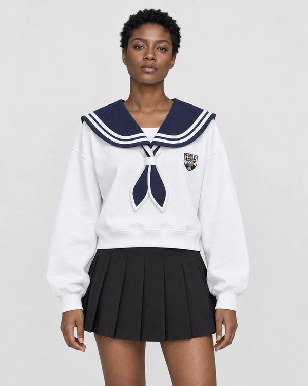 Woman in a cropped white sailor-collar sweatshirt with a navy collar and tie over a black pleated skirt, torso to mid-thigh