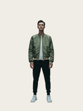 Person wearing a green bomber jacket, white shirt, black pants, and white sneakers on a light gray background