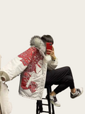 Person wearing a white jacket with red patterns and fur hood, sitting on a stool and using a phone.