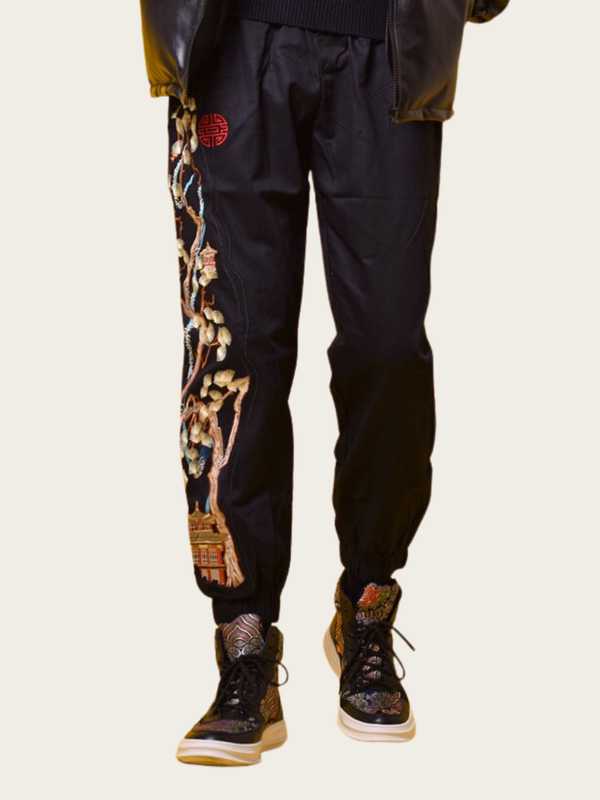 Black pants with floral embroidery and a red emblem worn by a person on a beige background
