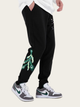 Black jogger pants with green peacock feather design worn by a person on a beige background