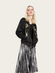 Woman wearing a black floral embroidered jacket and silver pleated skirt on a white background