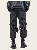 Person wearing black tactical pants with multiple pockets and straps on a beige background