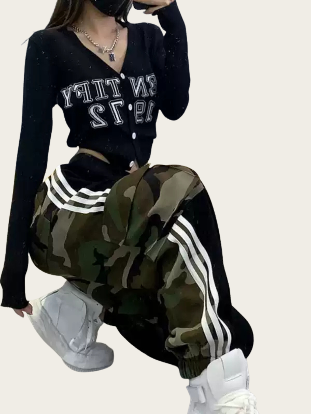 Person wearing a black cardigan with lettering, camouflage pants with white stripes, and white sneakers on a beige background.