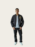 Person wearing a black bomber jacket with a logo on a beige background