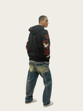 Person wearing a black hoodie with red designs and blue jeans on a white background