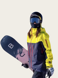 Person wearing a yellow and gray jacket with a blue helmet and goggles, holding a snowboard.