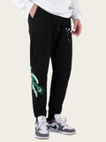 Person wearing black sweatpants with green peacock design and white sneakers on a light background
