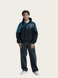 Person wearing a black hoodie with blue patterns and dark pants on a beige background