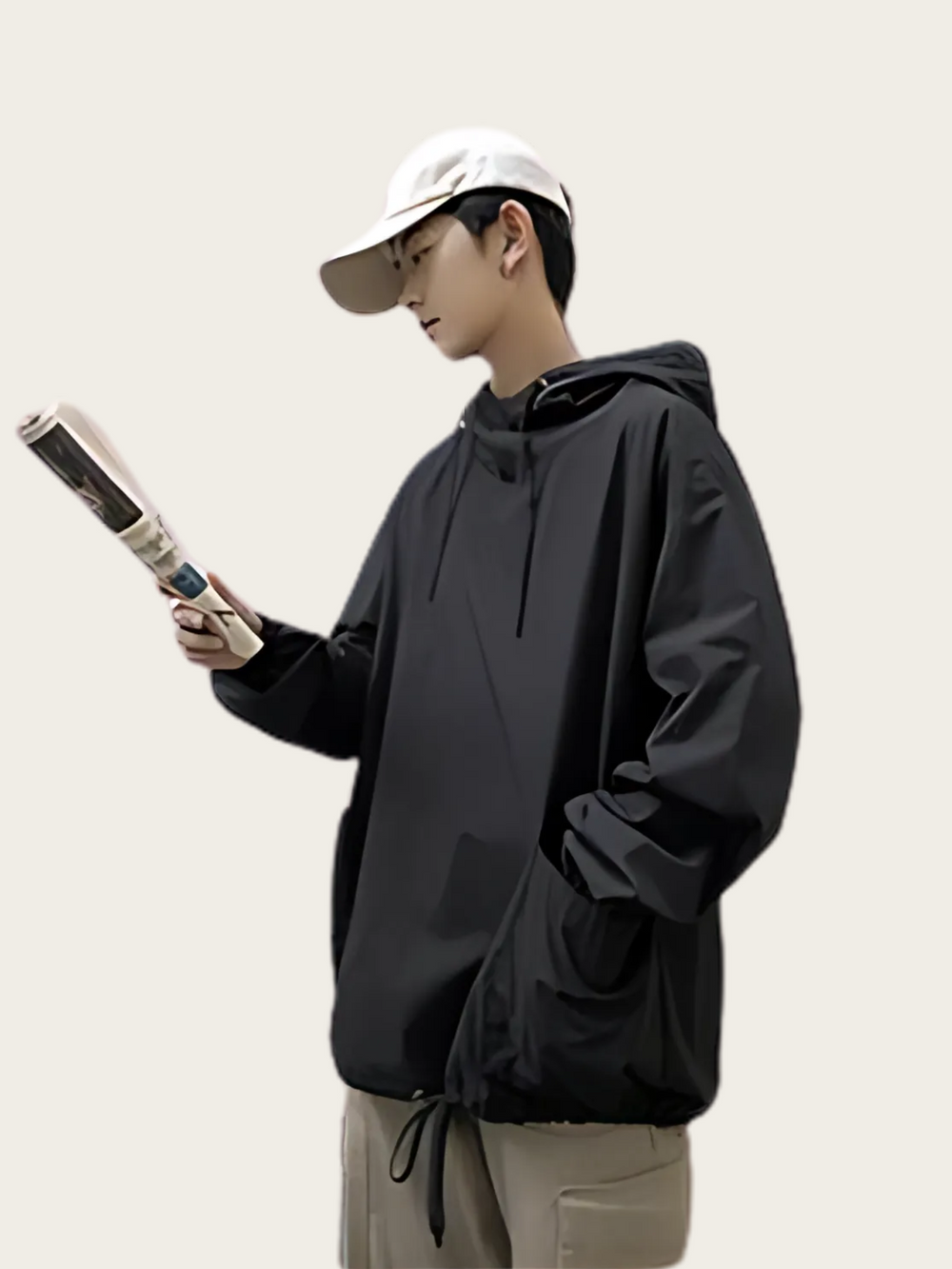Person wearing a black hoodie and beige cap holding a phone on a plain background
