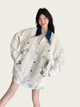 Woman wearing a white jacket with floral embroidery on a plain background