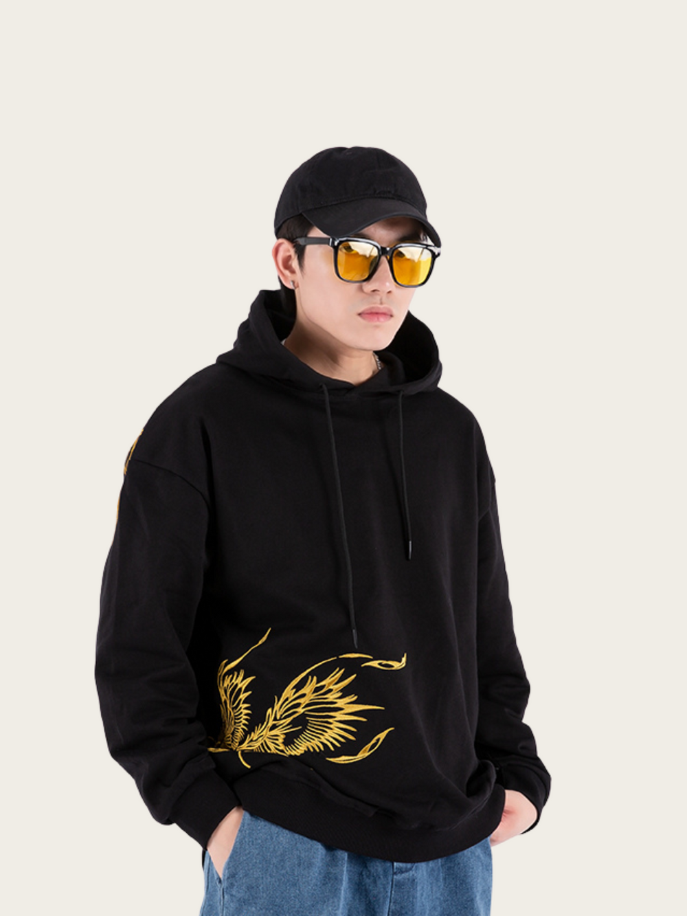 Person wearing a black hoodie with gold design, blue jeans, and sunglasses on a light gray background