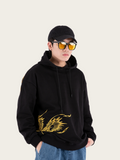 Person wearing a black hoodie with gold design, blue jeans, and sunglasses on a light gray background
