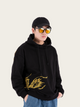 Person wearing a black hoodie with gold design, blue jeans, and sunglasses on a light gray background