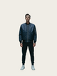 Person wearing a dark blue bomber jacket and black pants on a light beige background
