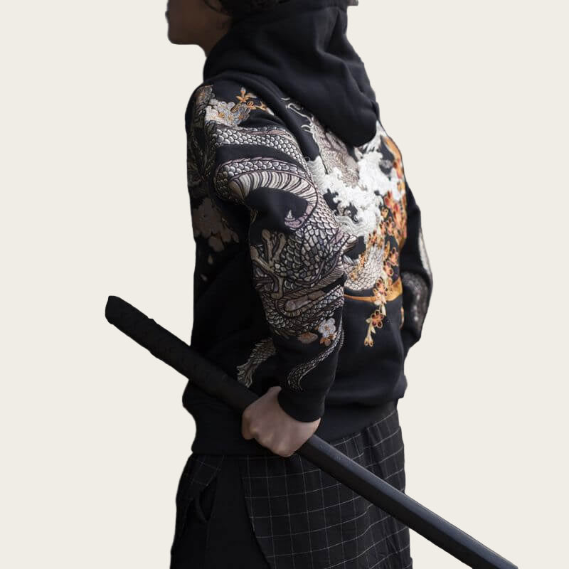 Person wearing a black hoodie with a colorful dragon design, standing against a metallic fence.