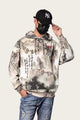 Person wearing a camouflage hoodie with text and graphics, blue jeans, and a black cap with a white logo.