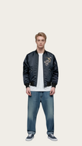 Man wearing a black bomber jacket with floral embroidery, white t-shirt, and blue jeans on a gray background