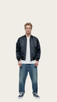 Man wearing a black bomber jacket, white shirt, and blue jeans on a gray background
