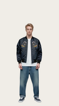 Man wearing a black bomber jacket with yellow designs, white shirt, blue jeans, and black shoes on a gray background