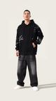 Person wearing a black hoodie with white patterns and dark jeans on a light background