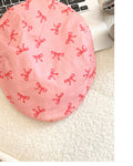 Top view of the pink flat cap with small red bows printed across the rounded crown, on a white knit blanket