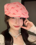 Woman wearing a pink flat cap with a scattered red bow print, one hand raised to her cheek, front view