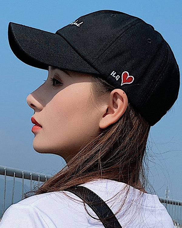 Woman wearing the black cap with white cursive script and a small red heart on the side, profile view