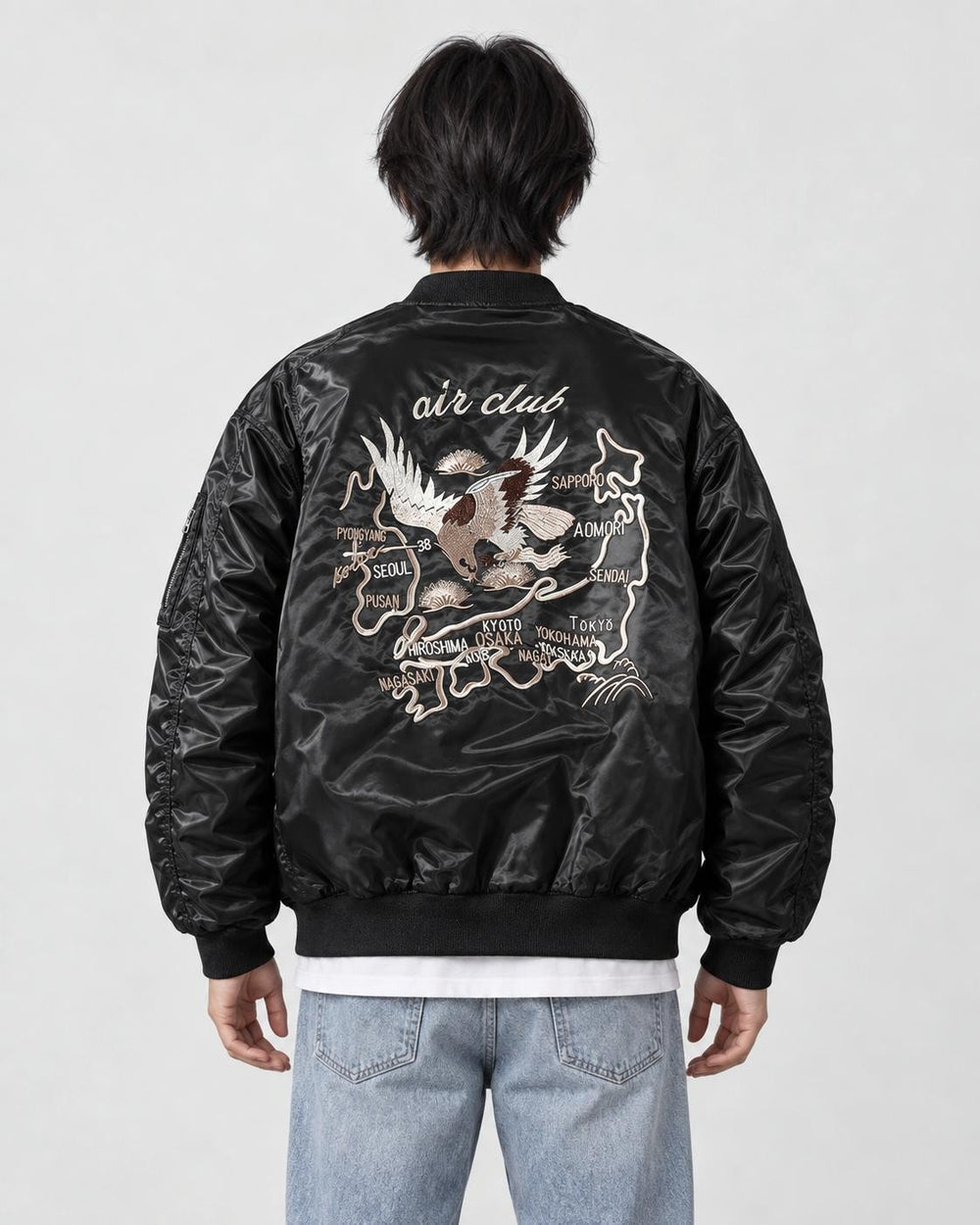 Model seen from behind in the black padded bomber jacket with a cream eagle and map embroidery, a white tee and light jeans