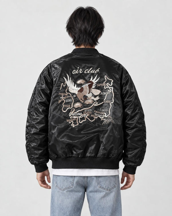 Model seen from behind in the black padded bomber jacket with a cream eagle and map embroidery, a white tee and light jeans