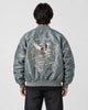 Model seen from behind in the slate gray padded bomber jacket with a cream eagle and map embroidery and black jeans