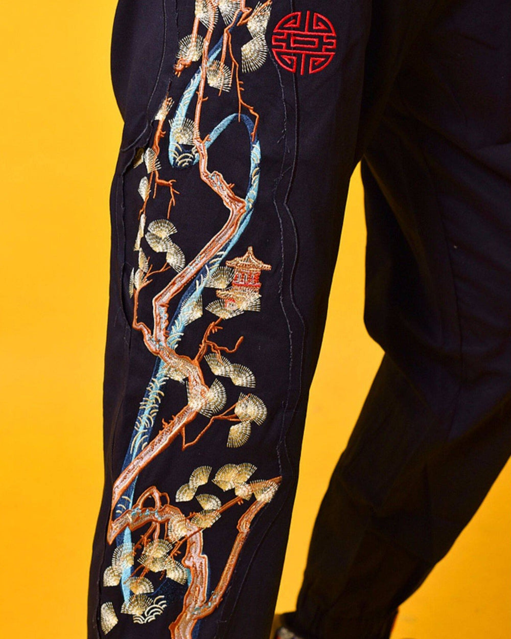 Close view of the embroidered pine tree in cream, pale green and copper thread with the red seal on the black joggers