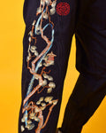 Close view of the embroidered pine tree in cream, pale green and copper thread with the red seal on the black joggers