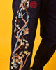 Close view of the embroidered pine tree in cream, pale green and copper thread with the red seal on the black joggers