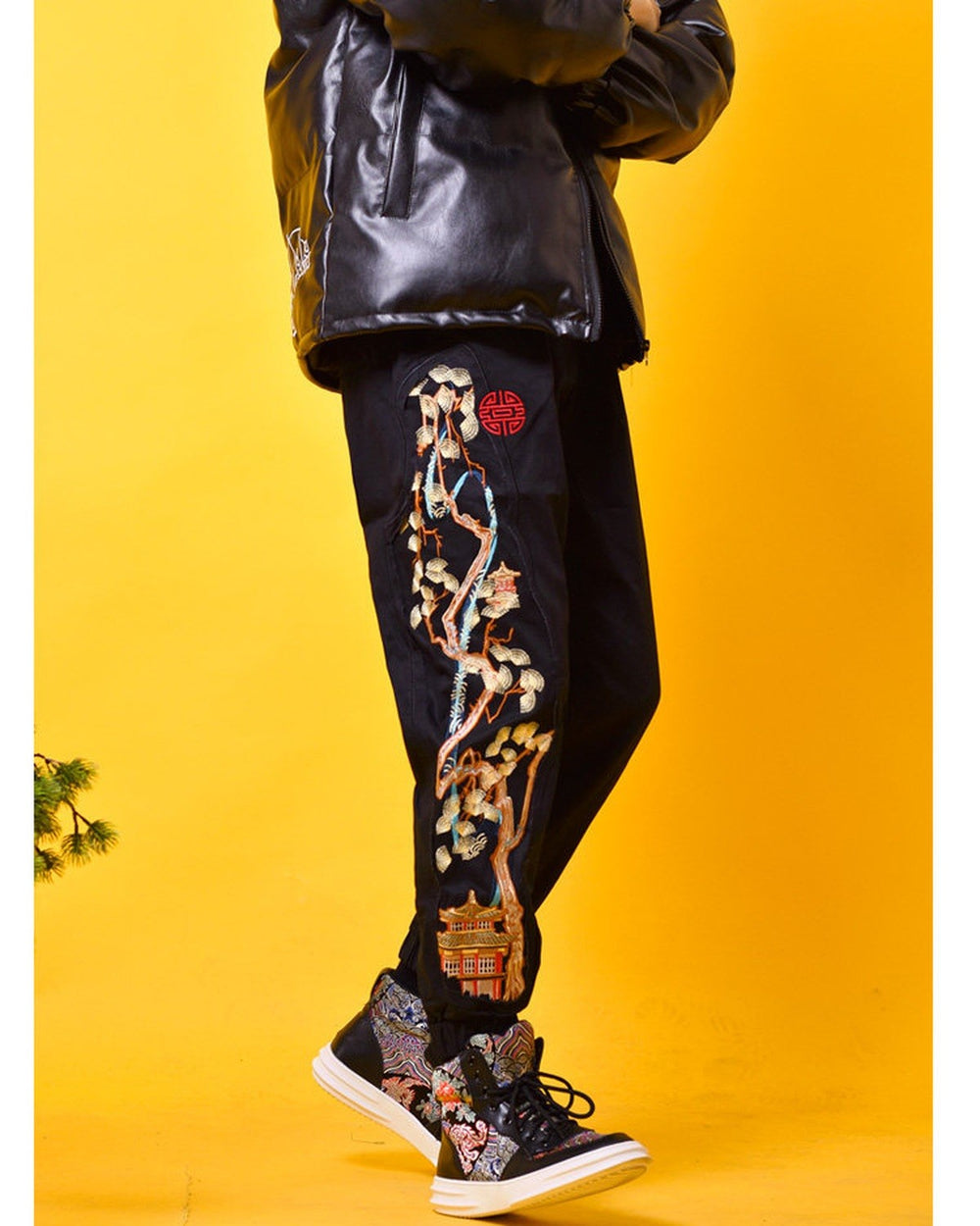 Black jogger pants with a pine tree and pavilion embroidered down the outer leg, side view of a model walking