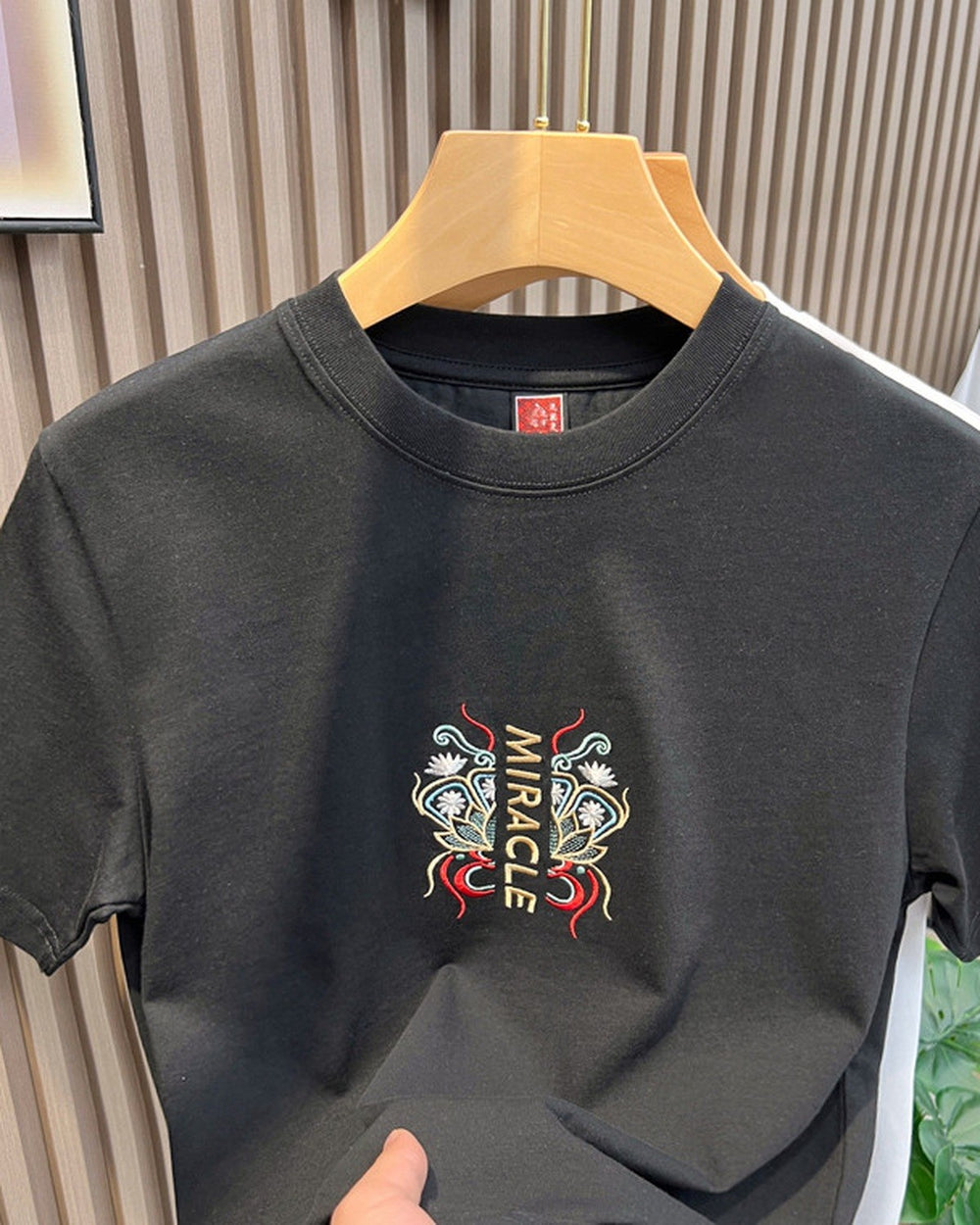 Front of the black T-shirt with a small embroidered MIRACLE emblem at the chest and a red label at the neck