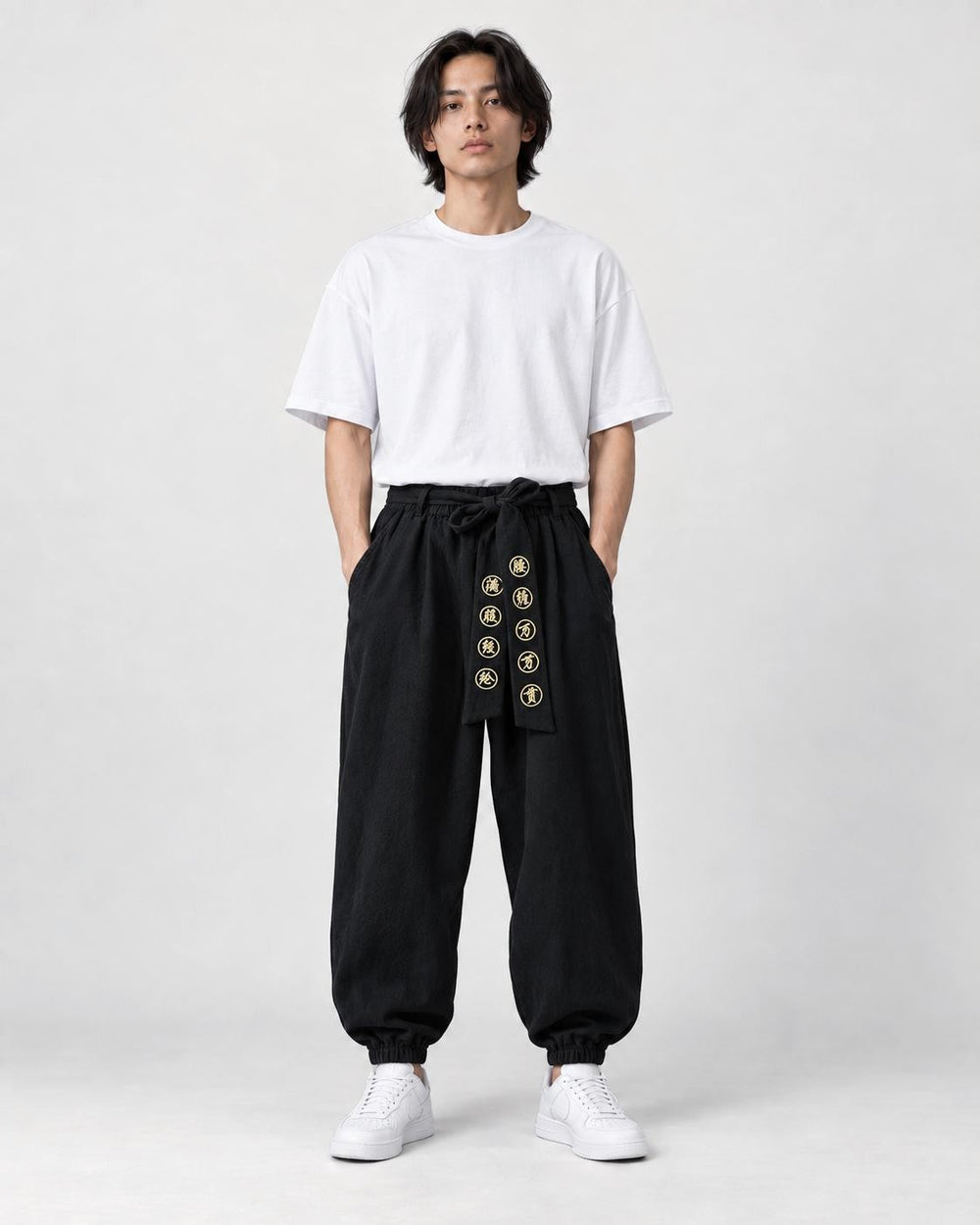 Model wearing black harem pants with a gold-embroidered sash, a white tee and white sneakers, front view