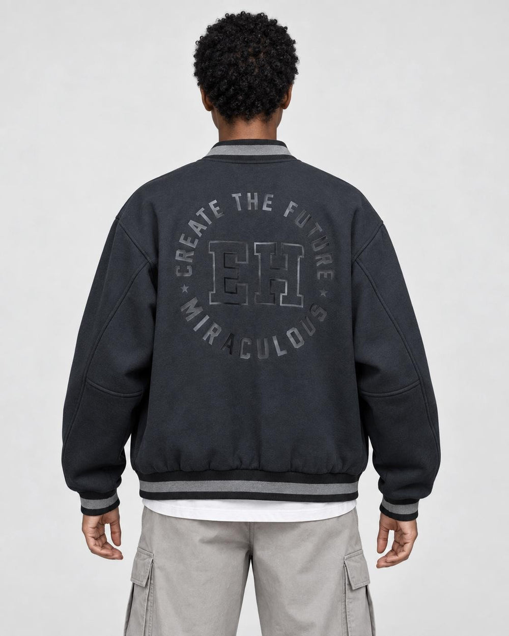 Model wearing the dark varsity jacket seen from behind, with the circular CREATE THE FUTURE, EH, MIRACULOUS print on the back