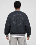 Model wearing the dark varsity jacket seen from behind, with the circular CREATE THE FUTURE, EH, MIRACULOUS print on the back