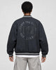 Model wearing the dark varsity jacket seen from behind, with the circular CREATE THE FUTURE, EH, MIRACULOUS print on the back
