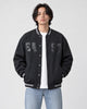 Model wearing the dark slate-black fleece-lined varsity jacket open over a white tee and light-wash jeans, front view