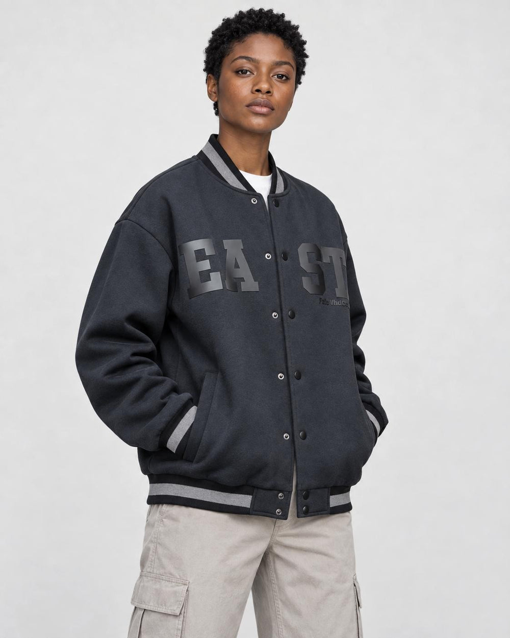Model wearing the dark varsity jacket with the snaps closed over a white tee and stone trousers, three-quarter view