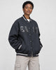 Model wearing the dark varsity jacket with the snaps closed over a white tee and stone trousers, three-quarter view